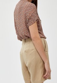 Blusa corta a maniche corte con motivo marrone abbinata a pantaloni corti in lino beige caratterizzati da una texture liscia e tasche posteriori. La blusa è arricciata in vita.