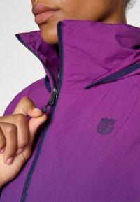 Lilla jakke med høy krage, har glidelås og marineblå detaljer. En liten brodert logo er synlig øverst til venstre. Glatt tekstur.
