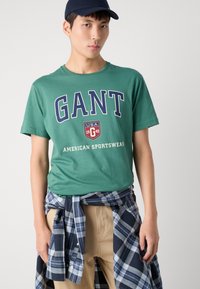Zelené bavlněné tričko s logem "GANT" v modré barvě. Kombinujte s khaki kalhotami a uvázanou námořnickou košile v kostkované úpravě pro vrstvený vzhled.