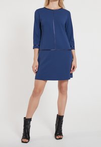 Marinblå klänning med trekvartslånga ärmar, utrustad med ett dragkedjedetalj och satin accent vid halsringningen och fållen. Bärs med svarta ankelstövlar.