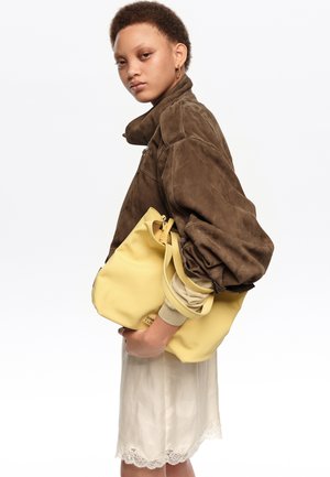 Joven con chaqueta marrón y falda color crema sosteniendo un gran bolso amarillo sobre un fondo blanco liso.