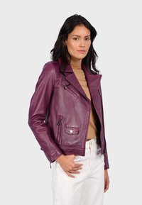 Burgundská kožená bunda s přiléhavým střihem, výstřihem do V, asymetrickým zipem a dvěma předními kapsami. Nositá přes béžový top.