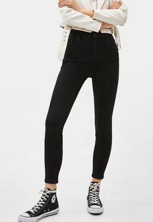 Person iført sorte skinny jeans, sorte høje sneakers og en hvid jakke med armene krydset, stående mod en hvid baggrund.