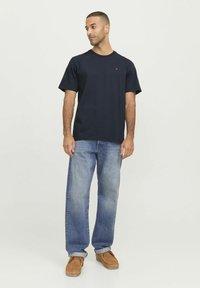 Camiseta de algodón de color navy con mangas cortas y escote redondo, que presenta un pequeño acento rojo en el pecho; combinada con jeans de mezclilla azul claro y calzado marrón.