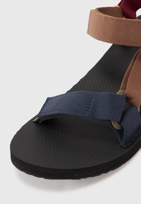 Sandalia multicolor con una suela de goma negra texturizada. Presenta tiras de tela en azul marino y marrón con un acabado suave y acentos en amarillo.