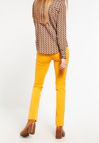 Blusa de manga larga con estampado geométrico en marrón, naranja y crema, combinada con pantalones ajustados amarillos y botines de tacón marrones.