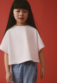 Haut blanc à manches courtes avec un col rond, associé à un pantalon en denim bleu clair à coupe décontractée avec une taille à cordon. Fond rouge.