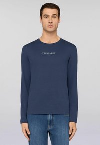 Mężczyzna w granatowej koszuli z długim rękawem z napisem "TRUSSARDI 1911", zestawionej z niebieskimi dżinsami, na jednolitym jasnoszarym tle.