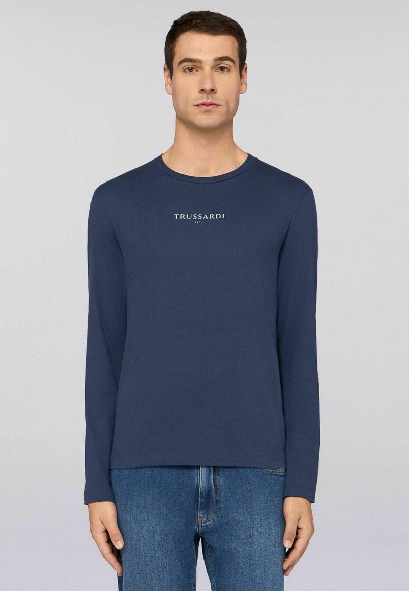 Mężczyzna w granatowej koszuli z długim rękawem z napisem "TRUSSARDI 1911", zestawionej z niebieskimi dżinsami, na jednolitym jasnoszarym tle.
