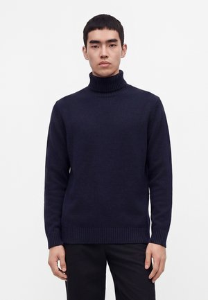 Homme de face portant un pull à col roulé côtelé bleu marine foncé et un pantalon noir sur un fond clair uni.