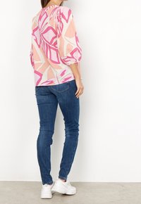 Blusa a maniche corte color rosa pastello con forme geometrice audaci in rosa e bianco, abbinata a jeans slim-fit blu scuro e sneakers bianche.