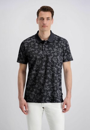 Man die een zwart poloshirt met bloemenpatroon en een witte broek draagt, staand tegen een effen lichte achtergrond.