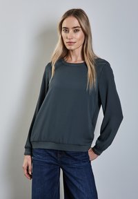 Mörkgrå långärmad topp med rund halsringning och elastisk nederkant. Tyget är slätt med en något lös passform. Blå denimbyxor med vida ben.
