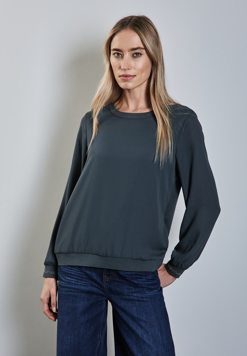 Mörkgrå långärmad topp med rund halsringning och elastisk nederkant. Tyget är slätt med en något lös passform. Blå denimbyxor med vida ben.