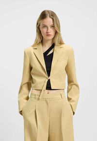 Geknipte gele blazer met een striksluiting, reverskraag en lange mouwen, gedragen over een zwart top met een asymmetrische snit.