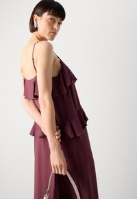 Bordeauxfarbene ärmellose Bluse mit Rüschen, aus strukturiertem Stoff. Das Model hält eine hellfarbige Handtasche und hat einen kurzen Haarschnitt.