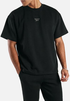Man draagt een zwarte korte mouwen Reebok t-shirt met klein logo op de borst en bijpassende zwarte broek, handen deels in de zakken.