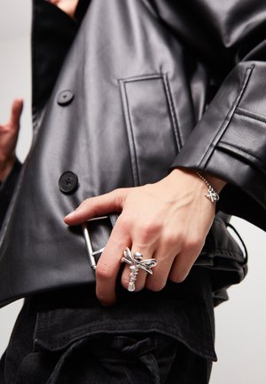 Chaqueta de cuero negro con grandes botones, hebilla de cinturón plateada y una mano con un anillo de plata texturizada con un diseño intrincado.