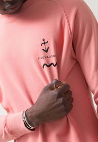 Sudadera rosa claro hecha de una tela suave, con gráficos de ancla negra y olas en el pecho. Cuello y puños acanalados visibles.
