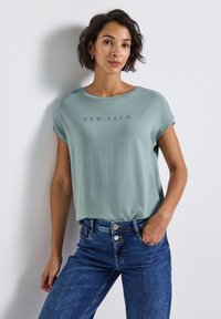 Mintgroene top met korte mouwen en een ronde hals, voorzien van de tekst "NEW PATH" in een donkerdere tint gedrukt. Combinatie met denim jeans.