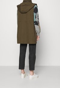 Olijfgroene mouwloze jas met een capuchon, over een grijze sweatshirt met een print. Gecombineerd met zwarte jeans en witte sneakers.