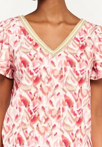 Top s výstřihem do V z lehké látky s růžovo-béžovým abstraktním vzorem; má zlatý lem podél výstřihu a krátkých rukávů.