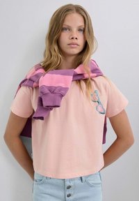 Camiseta de algodón rosa con mangas cortas, que presenta un pequeño bolsillo y un gráfico en azul y rosa. Un sudadera a rayas morada y rosa cae sobre los hombros.