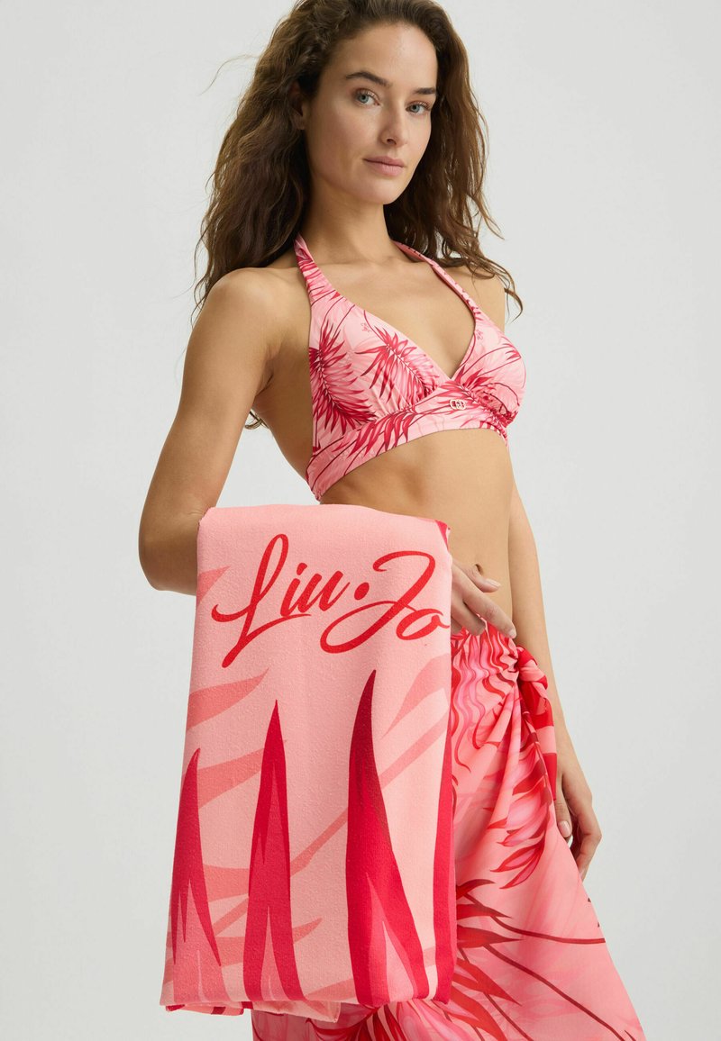 Bikini halter rosa con motivo foglia di palma abbinato a un asciugamano rosa con logo in rosso. La pelle è liscia, i capelli sono disordinati.