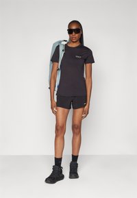 Czarny t-shirt i szorty w zestawie z jasnoniebieskim plecakiem. Ciemne okulary przeciwsłoneczne, solidne czarne buty trekkingowe i skarpetki do kostek dopełniają wygląd.
