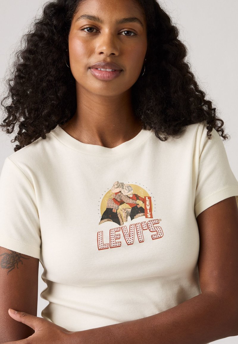 T-shirt en coton crème à manches courtes avec un graphisme de style rétro, "LEVI'S," et deux cowboys sur le devant. Texture lisse.
