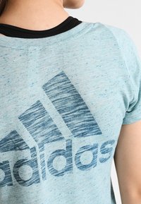 Valkoinen sininen urheilupaita, jossa on teksturoitu pinta ja suuri tummansininen Adidas-logo takana. Pyöreä kaula-aukko.