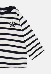 Camisa de manga longa com riscas em azul marinho e branco, com textura canelada e um logótipo de barco à vela no peito. Gola redonda com detalhe em botão.