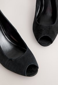 Czarne zamszowe buty na wysokim obcasie z otwartym noskiem, gładkim wnętrzem i zaokrąglonym obcasem. Charakteryzują się elegancką sylwetką i subtelną fakturą.