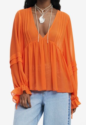 Vrouw draagt een doorschijnende oranje blouse met lange mouwen, plooien en gerimpelde manchetten, gecombineerd met lichtblauwe spijkerbroek en gelaagde zilveren kettingen.