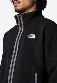 Svart fleecetröja med hög krage, med en fullständig framdragkedja och en vit logotyp på bröstet. Synlig sömnadsdetaljer.