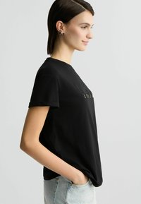 Lühikeste varrukatega must särk pehmest kangast, ümmarguse kaelusega ja minimalistliku disainiga, mille esiküljel on kuldsed aktsendid.