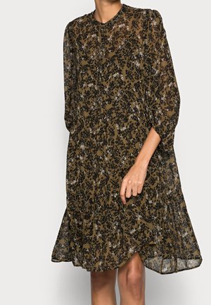 Brun gennemsigtig midi-kjole med sort blomsterprint, lange puffede ærmer og en flydende A-formet silhuet på en stående person.