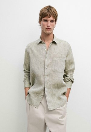 Junger Mann trägt ein hellbeiges, langärmliges Hemd mit Knöpfen und einer Brusttasche, steht mit den Händen in den Taschen vor einem schlichten weißen Hintergrund.