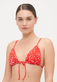 Haut de bikini rouge avec des bonnets triangulaires, motif floral blanc, bretelles ajustables et fermeture à nouer à l'avant. Texture de tissu lisse.