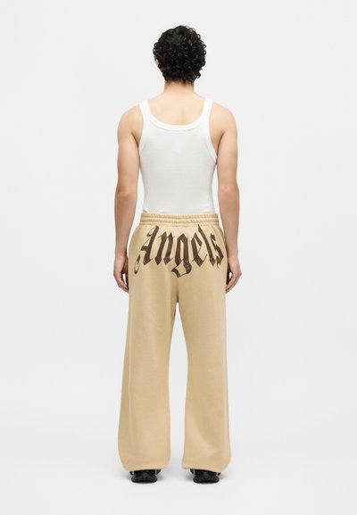Άνδρας με σγουρά μαλλιά, που φοράει λευκό αμάνικο μπλουζάκι και μπεζ παντελόνια με πλατύτερα πόδια με την επιγραφή «Angels» στην πλάτη, στέκεται με την πλάτη προς τα εμάς.