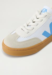 Tênis com parte superior em lona branca e destaques em camurça bege, apresentando um logótipo azul, sola de borracha tipo gum e atacadores redondos. Design clássico.