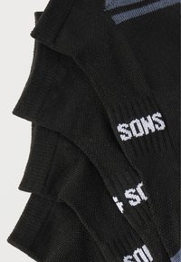 Quatre paires de chaussettes noires chevilles empilées, chacune avec un texte blanc partiellement visible près du bord-côte sur un fond uni.
