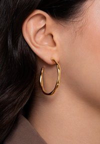 Orecchino a cerchio in oro con un design ondulato, caratterizzato da una superficie liscia e lucida. L'orecchino è aperto, formando una forma a U.