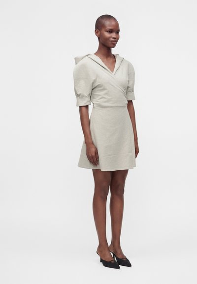 Femme portant une robe à capuche beige clair à manches courtes avec un devant croisé et des escarpins noirs pointus, debout devant un fond uni.