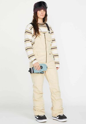 Mujer con mono de nieve beige, camiseta de manga larga a rayas, zapatillas negras, gorro morado, gafas de esquí en la cabeza, sosteniendo guantes azules.