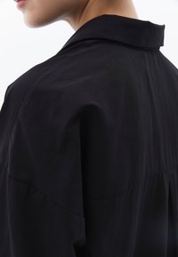 Zwarte shirt met een klassieke kraag en een ontspannen pasvorm. Zachte textuur met zichtbare stiksels en plooidetails aan de achterkant.