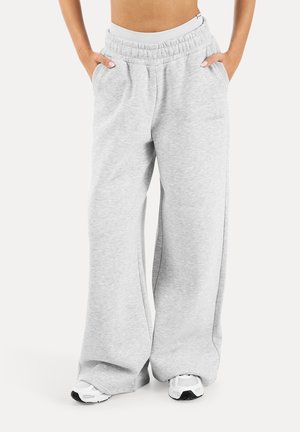 Person iført løse, lysegrå joggingbukser med elastisk talje og hvide sneakers, hænder i lommerne, stående mod en ensfarvet baggrund.