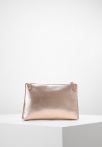 Bolso de mano metálico en oro rosa con cierre de cremallera y pequeños anillos dorados a cada lado, colocado sobre un pedestal blanco.