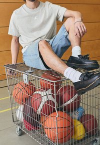 Šedé bavlněné tričko s logem, džíny šortky, černé boty a drátěný koš plný různých sportovních míčů, včetně basketbalových a fotbalových míčů.