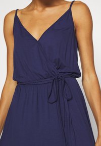 Femme portant une robe cache-coeur sans manches bleu marine avec de fines bretelles et une ceinture nouée à la taille, montrant le haut du torse et les bras.